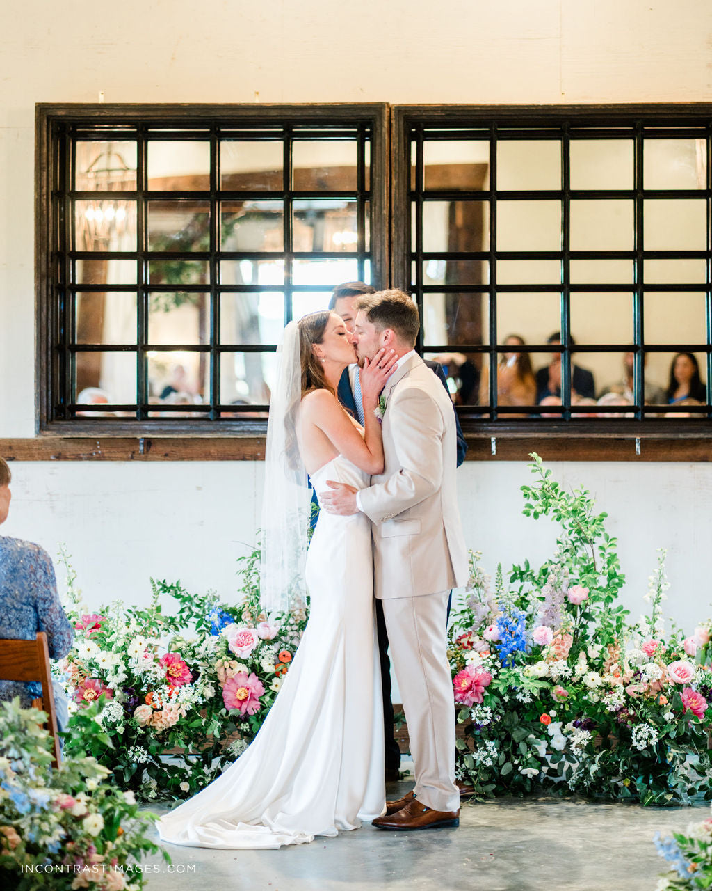 Couple embracing in a wedding setting with floral decorations and people in the background.