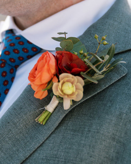 Man wearing a gray suit with a blue tie, orange pocket square, and floral boutonniere.